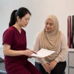 Chinese female clinician in maroon scrubs explaining back pain care options to a Malaysian patient in a clinic