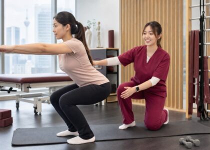 Female One Spine clinician in maroon scrubs coaching a Malaysian patient through a safe squat in a rehabilitation clinic
