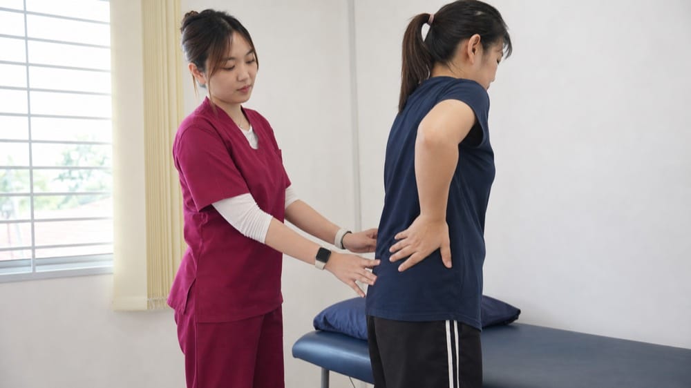 Ivy Ting from One Spine in maroon scrubs explaining lower back spasm assessment with a Malaysian patient