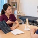 One Spine clinician in maroon scrubs explaining X-ray and MRI findings with a Malaysian patient in a clinic consultation
